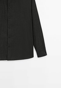 Chemise noire à manches longues boutonnée avec de fines rayures verticales et un ourlet incurvé sur fond blanc.