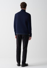 Muž otočený chrbtom, oblečený v tmavomodrom saká s pletenými rukávmi a golierom, čiernych nohaviciach a čiernych spoločenských topánkach na bielom pozadí.