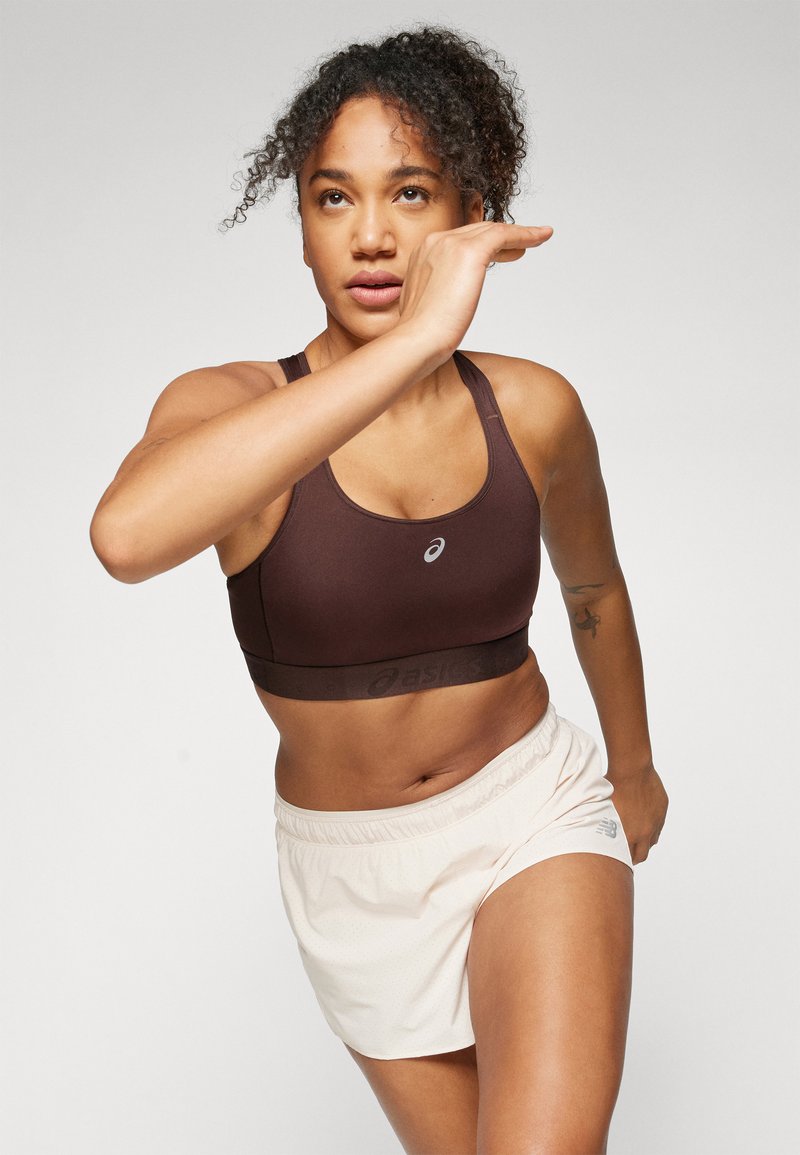 Braunensport-BH mit Logo, kombiniert mit hellbeigen Shorts mit einer strukturierten Gestaltung. Das Model befindet sich in einer Laufpose.