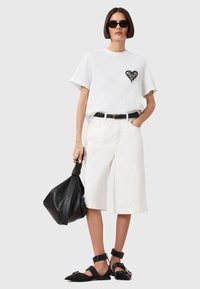Hvid bomulds t-shirt med sort hjerte-design, parret med hvide shorts til knæene, sort bælte og sort håndtaske. Model der bærer solbriller og flade sko.