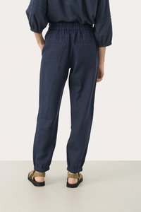 Pantaloni di lino blu navy con vita elasticizzata, gambe affusolate e polsini elasticizzati, abbinati a sandali verdi su una superficie neutra.