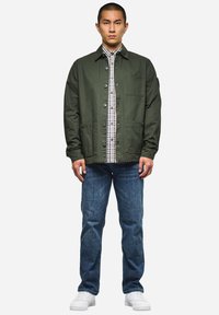 Olivgrüne Jacke mit Knopfverschluss und Brusttasche, über einem blau-weißen karierten Hemd getragen, kombiniert mit blauen Jeans und weißen Sneakers.