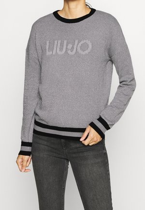 Kobieta ubrana w szary dzianinowy sweter z czarnym kołnierzem i mankietami, z napisem "LIU·JO" na piersi, w połączeniu z ciemnoszarymi jeansami, ręka oparta blisko pasa.