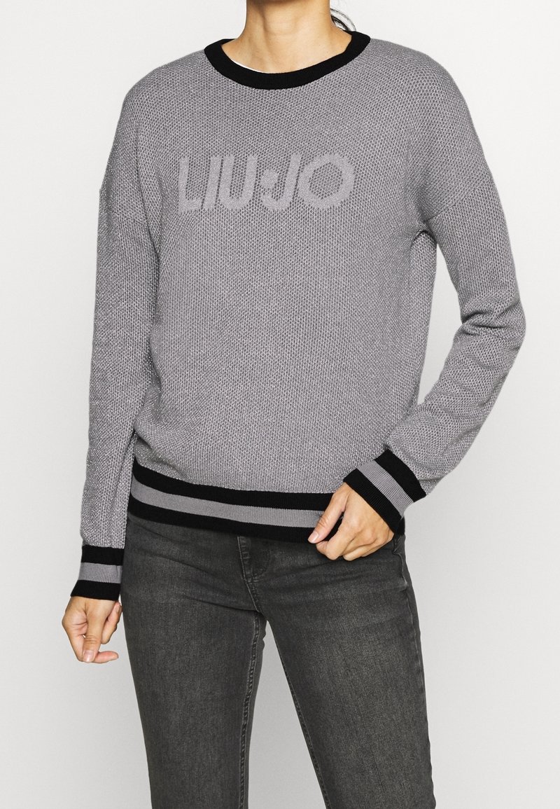 Femme portant un pull gris en maille avec col et poignets noirs, texte "LIU·JO" sur la poitrine, associée à un jean gris foncé, main reposant près de la taille.