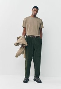 Beige póló, olívazöld bordás nadrág, és egy világos barna dzseki az egyik kezében. Barna, vastag talpú bakancsok.