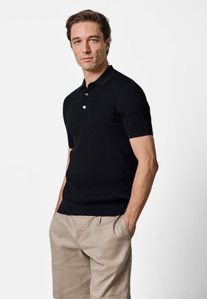 Mężczyzna stojący z rękami w kieszeniach, ubrany w czarną, krótką, dzianinową koszulkę polo i beżowe spodnie na jednolitym, jasnym tle.