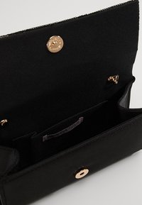 Svart rektangulär clutch-väska öppen för att visa tyginteriör, litet internt fack och guldmagnetisk knappstängning.