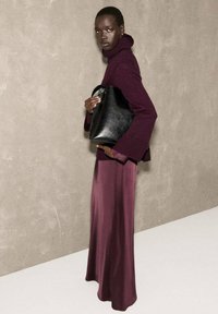 Maglione a dolcevita color bordeaux abbinato a una gonna viola fluida; la modella tiene in mano una borsa in pelle nera con una forma arrotondata e manico.