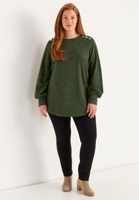 Groene gebreide trui met lange ballonmouwen, ronde hals en knoopaccenten op de schouders, gecombineerd met zwarte leggings en beige enkellaarsjes.