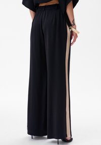 Zwarte ruime broek met een elastische tailleband en een beige zijstreep. De stof is glad en heeft een vloeiende textuur.