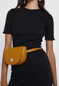 Musztardowożółta skórzana torba na ramię z zaokrągloną klapką, srebrnymi okuciami i dopasowanym regulowanym paskiem, noszona na czarnej prążkowanej sukience.