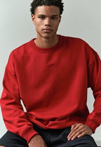 Rød sweatshirt lavet af blødt stof, med rund hals, sænkede skuldre og prægede "EDIT." logo på brystet. Mørke shorts fuldender looket.
