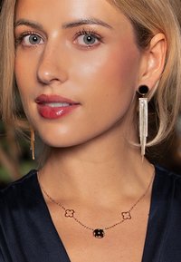 Gold-tone jewelry features a delicate chain with two floral accents and a round stone. Black circular earrings hang with thin fringe detailing.