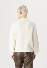 Weißer, flauschiger Strickpullover mit hohem Kragen und überschnittenen Schultern. Die Rückseite hat einen enganliegenden, gerippten Saum, kombiniert mit braunen Lederhosen.