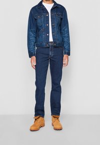 Chaqueta vaquera en azul oscuro, con cierre de botones y bolsillos, combinada con vaqueros azul oscuro y botas de ante marrón claro con cordones.