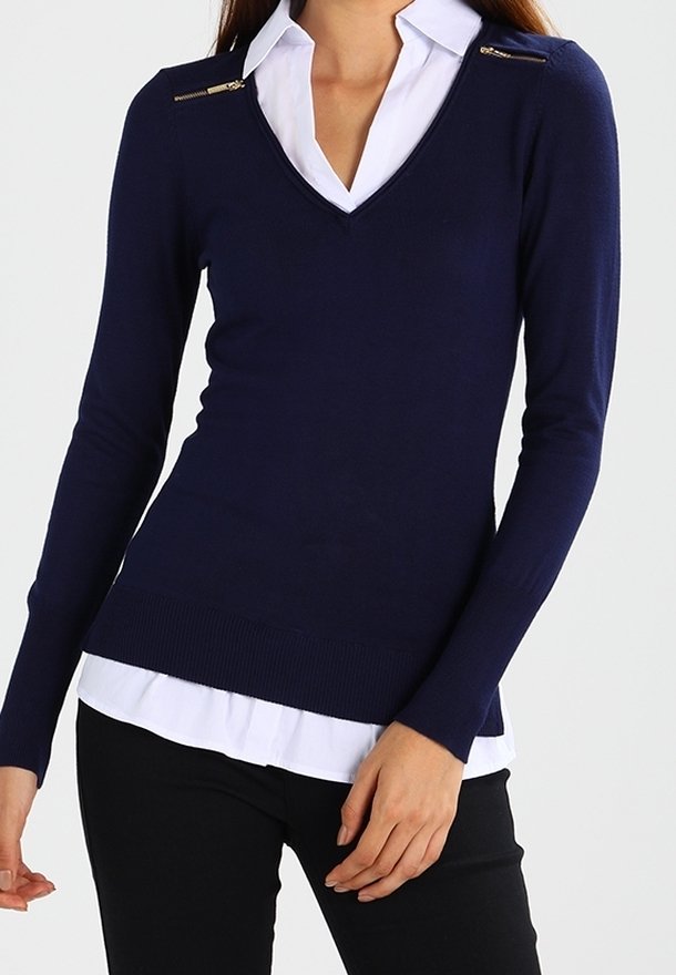 Marineblauwe gebreide trui met een V-hals en twee met rits afgesloten accenten bij de schouders, gecombineerd met een witte blouse met kraag en zwarte broek.