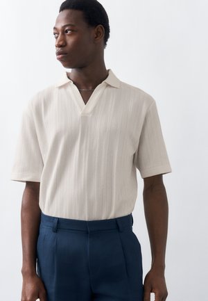 Mann mit kurzem Haar, der ein cremefarbenes, kurzärmliges Strick-Poloshirt trägt, das in marineblaue Hosen gesteckt ist, und seitlich vor einem weißen Hintergrund schaut.