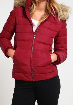 Kobieta w czerwonej pikowanej kurtce zapinanej na zamek z futrzanym kapturem i czarnych spodniach, z rękami w kieszeniach kurtki na jednolitym tle.