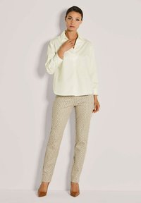 Frau in beigen, geometrisch gemusterten Hosen, cremefarbenem Langarmbluse und braunen spitzen Absätzen, die vor einem schlichten hellen Hintergrund steht.