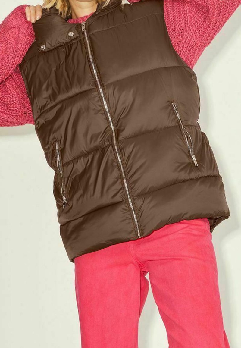 Gilet rembourré marron avec col haut, doté de deux poches zippées et d'un zip frontal, porté sur un pull rose et un pantalon rose.