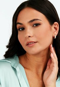 Nahaufnahme einer Person mit langen dunklen Haaren, die ein hellblaues Satintop und strukturierte Creolen trägt. Der Hauttyp ist warm mit natürlichem Make-up.