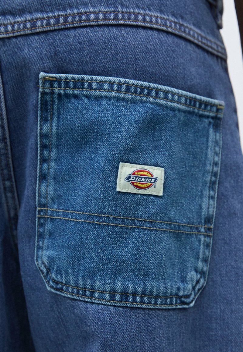 Denim broek met een achterzak voorzien van een genaaide Dickies logo label. De stof is medium blauw met zichtbare stiksels en een gladde textuur.