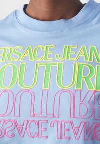 Světle modré bavlněné tričko s výrazným, vícbarevným textem: "VERSACE JEANS COUTURE" v žluté, zelené a růžové barvě s zrcadlovým efektem.