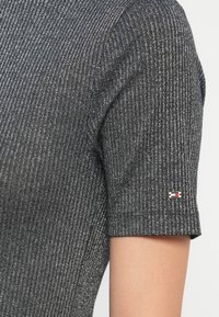 Tričko s krátkým rukávem vyrobené z texturované šedé látky s vertikálními pruhy. Na rukávu se nachází malá nášivka s logem v červené, bílé a modré barvě.