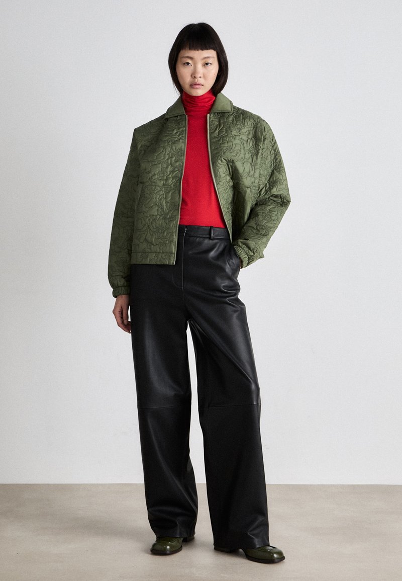 Quilted olivgrüner Bomberjacke mit einem schwarzen Reißverschluss, rotem Rollkragenpullover, schwarzen Fake-Leder weit geschnittenen Hosen und dunkelgrünen Schuhen.