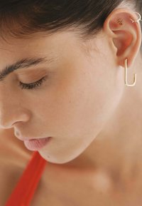 Pendientes de aro dorados y en forma de U, que muestran un diseño elegante. Piel texturizada con un suave enfoque en las características. Parte superior roja visible en el fondo.