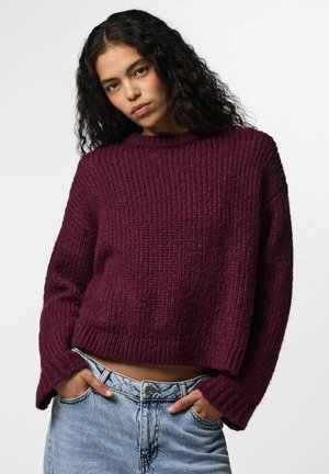 Ung kvinde med krøllet mørkt hår iført en løs vinrød strikket sweater og lyseblå jeans med hænderne i lommerne.