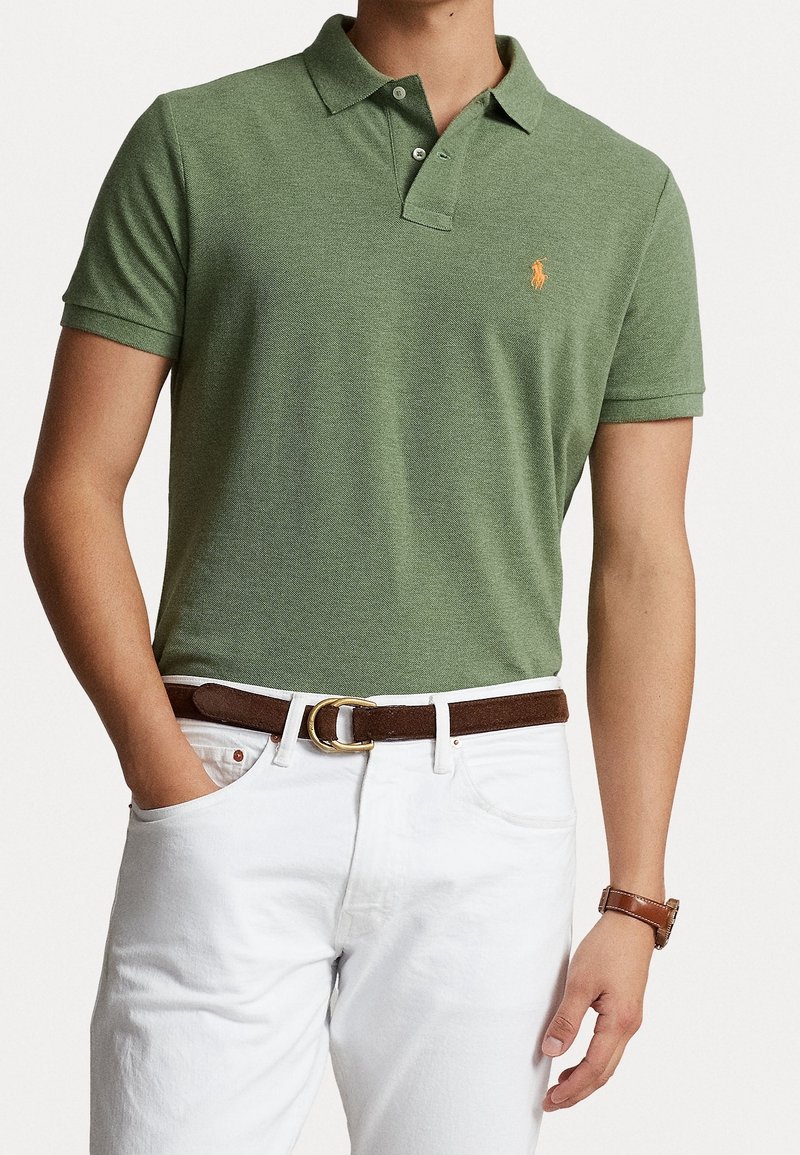 Homme portant un polo vert à manches courtes, un pantalon blanc avec une ceinture marron et une montre, une main dans la poche, vu du cou à la cuisse.