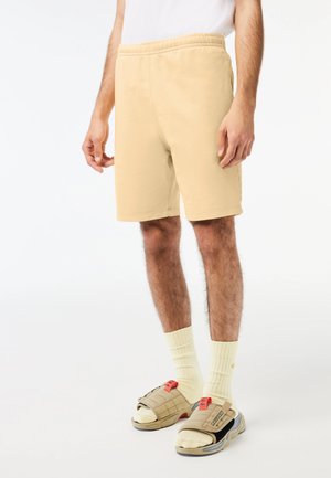 Shorts en coton beige avec une taille élastique, arrivant au genou, associés à des chaussettes jaunes à côtes et des sandales grises avec des accents rouges.