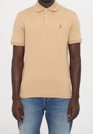 Mand iført beige kortærmet poloshirt med knapper og lille logo på brystet, kombineret med blå denimjeans.