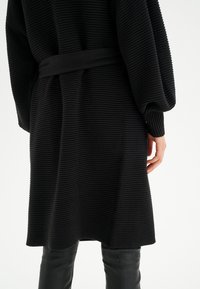 Mustat teksturoitu neuletakki, jossa on käsinukkasuunnittelu, vyötärölle sidottu vyö ja leveät hihat, yhdistettyinä tiukkoihin mustiin nahkahousuihin.