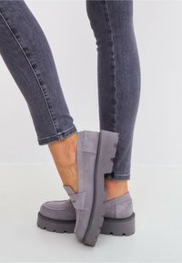 Grijze suede instappers met dikke profielzolen, gedragen met donkergrijze slim-fit jeans, getoond op gekruiste benen tegen een lichte achtergrond.