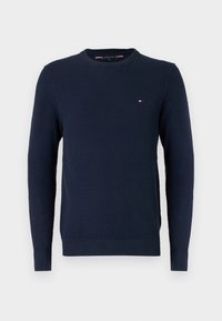 Tummansininen ribbiknitti crewneck-neulepusero, jossa on pitkät hihat ja pieni punainen, valkoinen ja sininen logo vasemmassa rinnassa.