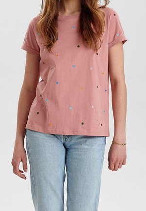 Mujer con camisa rosa de manga corta con lunares multicolores y vaqueros azul claro, de pie con los brazos relajados a los lados.