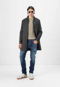 Manteau en mélange de laine gris avec une seule poche, pull en tricot beige, jeans bleus et baskets blanches avec des accents marron ; lunettes de soleil rondes.
