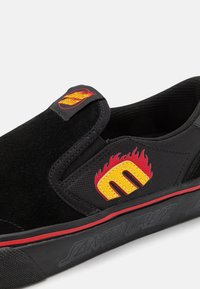 Melni slip-on apavi ar zamšādu, ar sarkanu un dzeltenu liesmu logotipu, un gumijas zoli ar sarkanu apdari un iegrieztu dizainu.