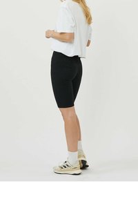 Shorts de vélo noirs avec une texture lisse, un design ajusté et une longueur jusqu'au mi-cuisse, associés à une chemise blanche ample et des baskets beiges.