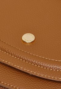 Sac à main en cuir texturé marron avec un emblème circulaire en or portant l'inscription "Joules" embossée. Détails de couture le long des bords.