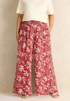 Person iført viddebenede, røde blomstrede bukser med hvid elastisk talje og en hvid kortærmet top, stående med den ene hånd i lommen.