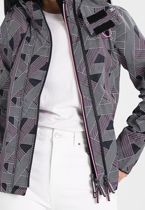 Zwart-witte jas met geometrisch patroon en roze rits, gedragen over een wit shirt en een witte broek.