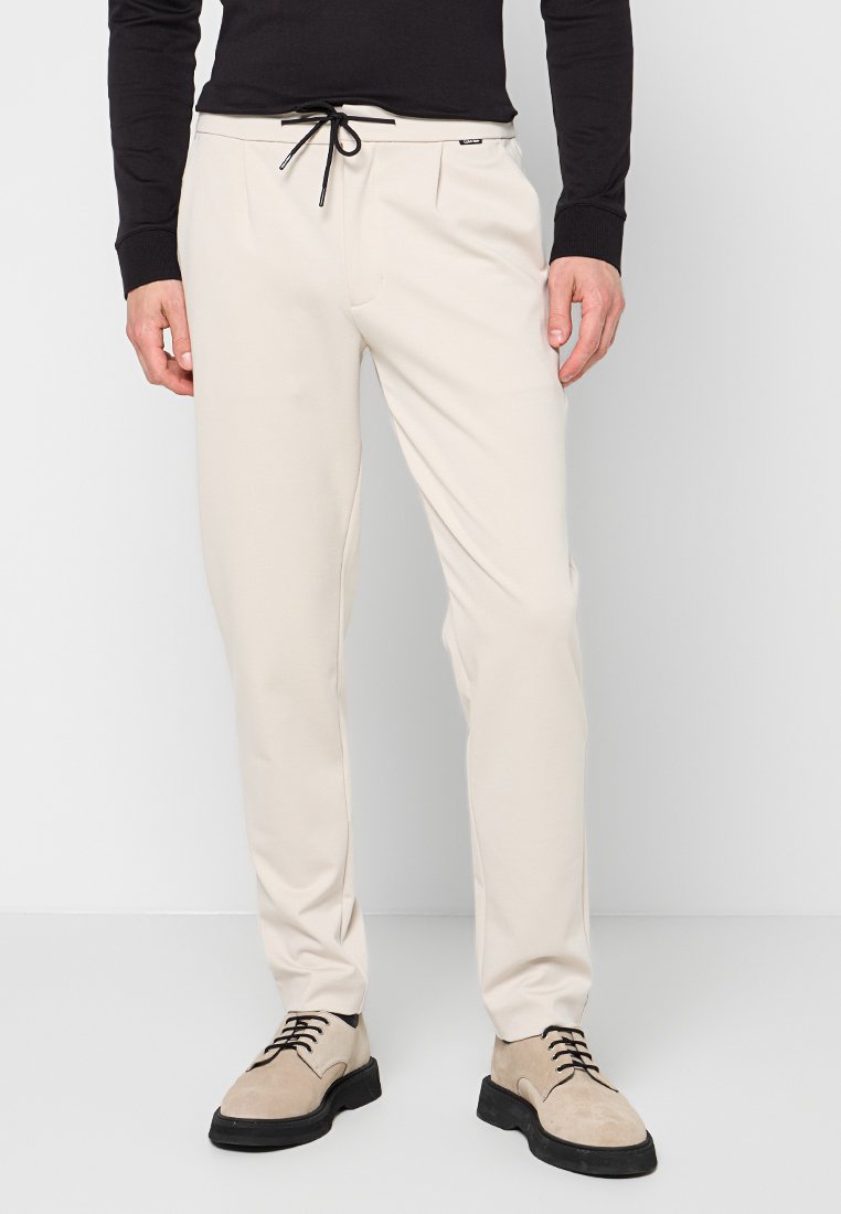 Beige maßgeschneiderte Hose mit elastischem Bund, zwei Fronttaschen und glatter Textur. Getragen mit hellbeigen Schnürschuhen.