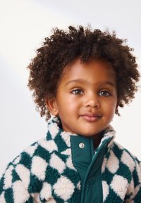 Bambino che indossa una giacca in pile a quadretti verde e bianca con colletto alto e chiusura a scatto. Capelli ricci, sfondo neutro.