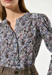 Blumen Bluse mit strukturiertem Muster in Blau-, Lila- und Grüntönen, mit Knopfkragen und puffärmel. Kombiniert mit einem karierten Taillenbund.
