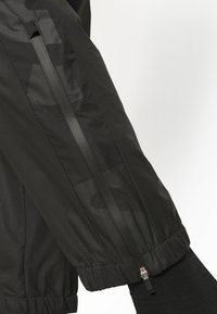 Černé sportovní kalhoty z hladkého, lehkého materiálu. Obsahují skrytý zip podél bočního švu a elastické manžety na kotnících.