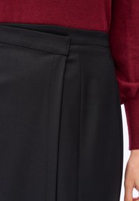 Close-up van een persoon in een zwarte wikkelrok en een rood langemouwen top, met zichtbare stoftextuur en details aan de zijkant.