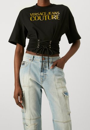 Czarny krótki T-shirt z złotym napisem "Versace Jeans Couture" i detalem sznurowania, zestawiony z jasnoniebieskimi jeansami cargo z kieszeniami.